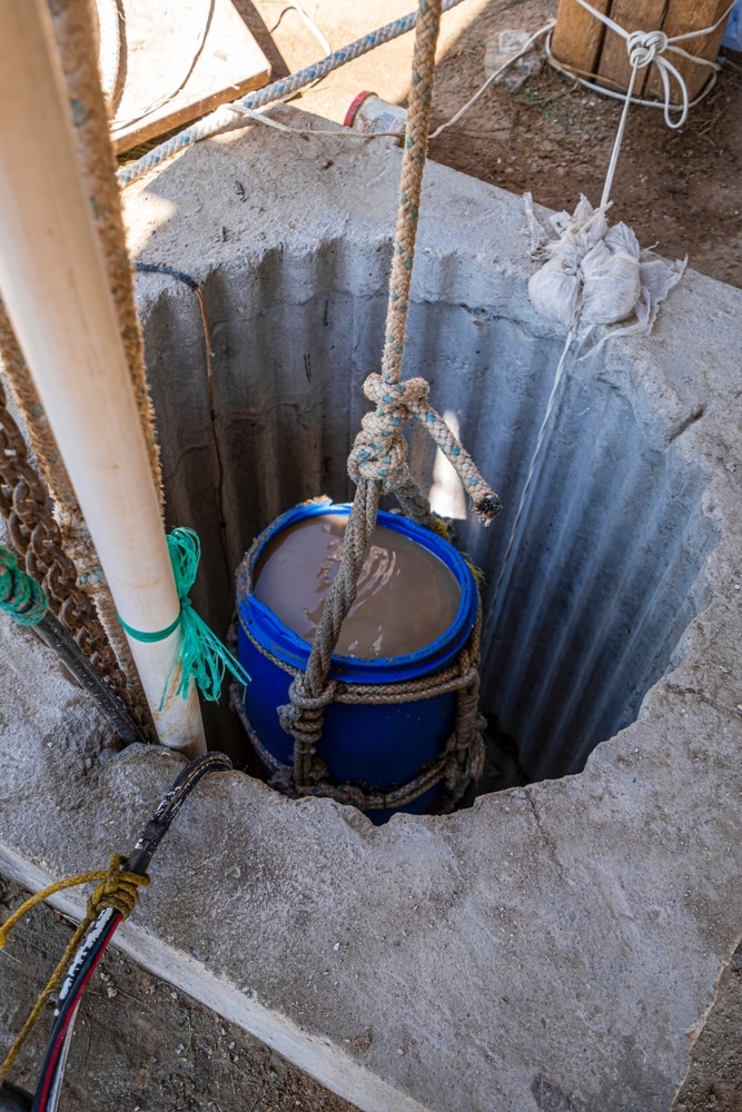Forage de puits : extraction d'eau boueuse au baril bleu Baril bleu rempli d'eau boueuse suspendu par des cordes dans un puits en béton lors d'un chantier.