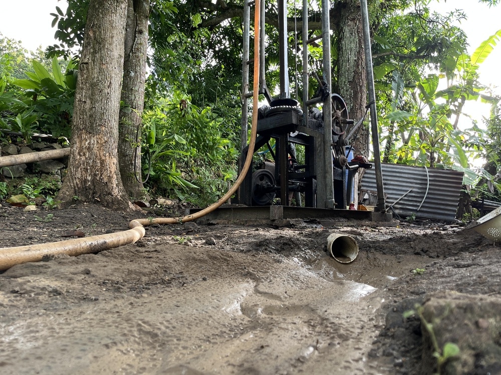 Forage de puits : machine et installation en extérieur Foreuse mécanique de puits dans un terrain boueux entouré d'arbres tropicaux et d'un tuyau orange.